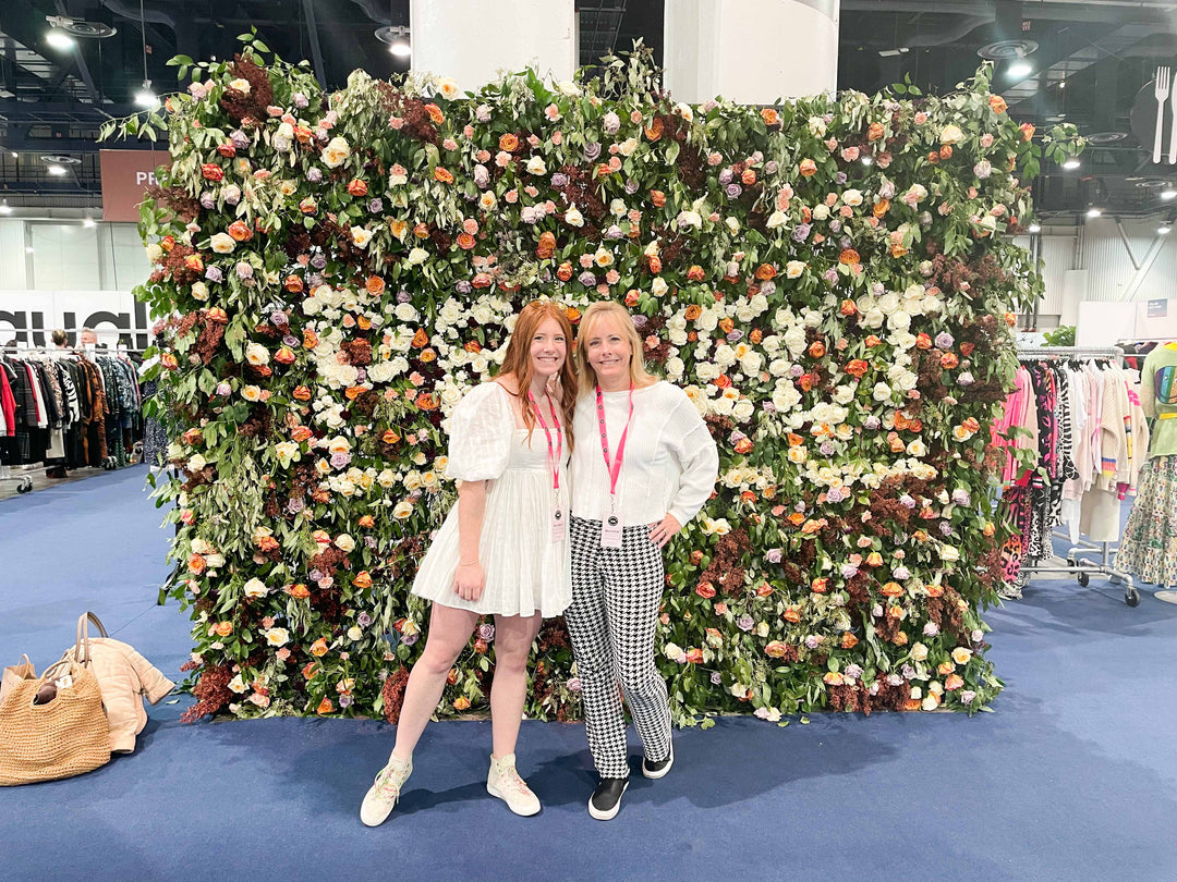 Two women standing in front of a beautiful flower wall at a convention. The woman on the left is wearing a white babydoll dress, while the woman on the right is dressed in houndstooth knit pants and a basic white long sleeve top. They are both smiling and